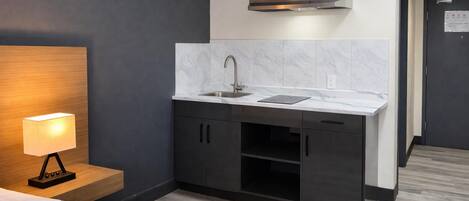 A compact hotel room with a bed, a bedside lamp, a kitchenette with a sink, cabinets, and a stove hood.