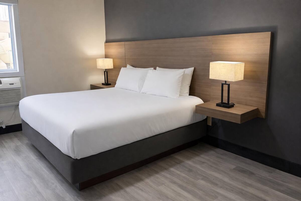 Une chambre d’hôtel avec un grand lit, deux tables de chevet avec des lampes, une tête de lit sombre et une fenêtre avec un appareil de climatisation.