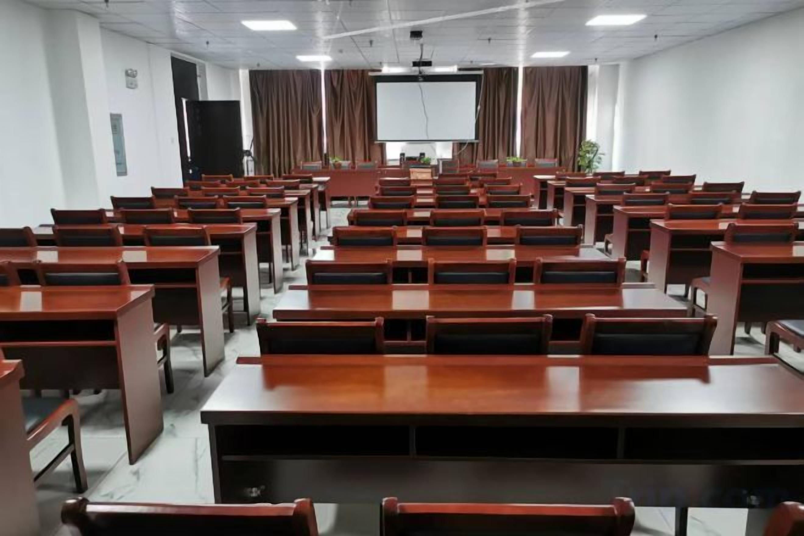 Un salón de conferencias con varias filas de mesas y sillas, una pantalla de proyector y una pizarra.