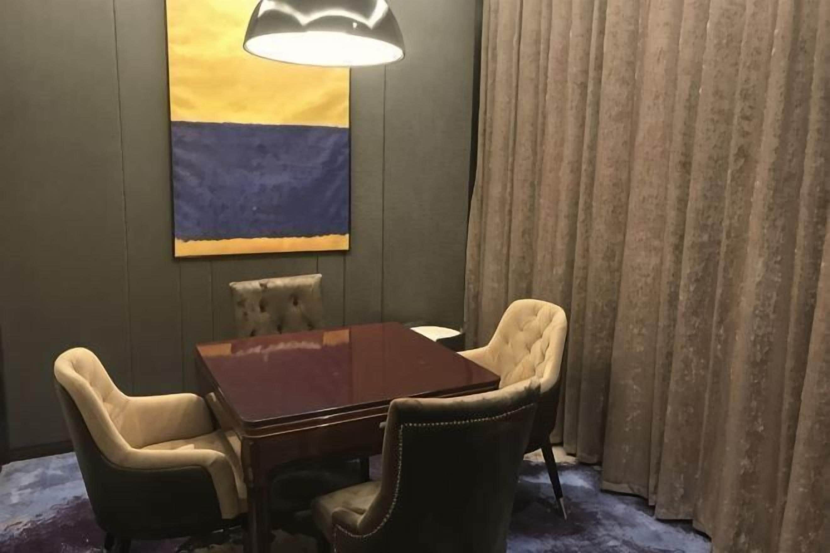 A room with a wooden table, four chairs, a lamp, a large abstract painting, and a curtain.