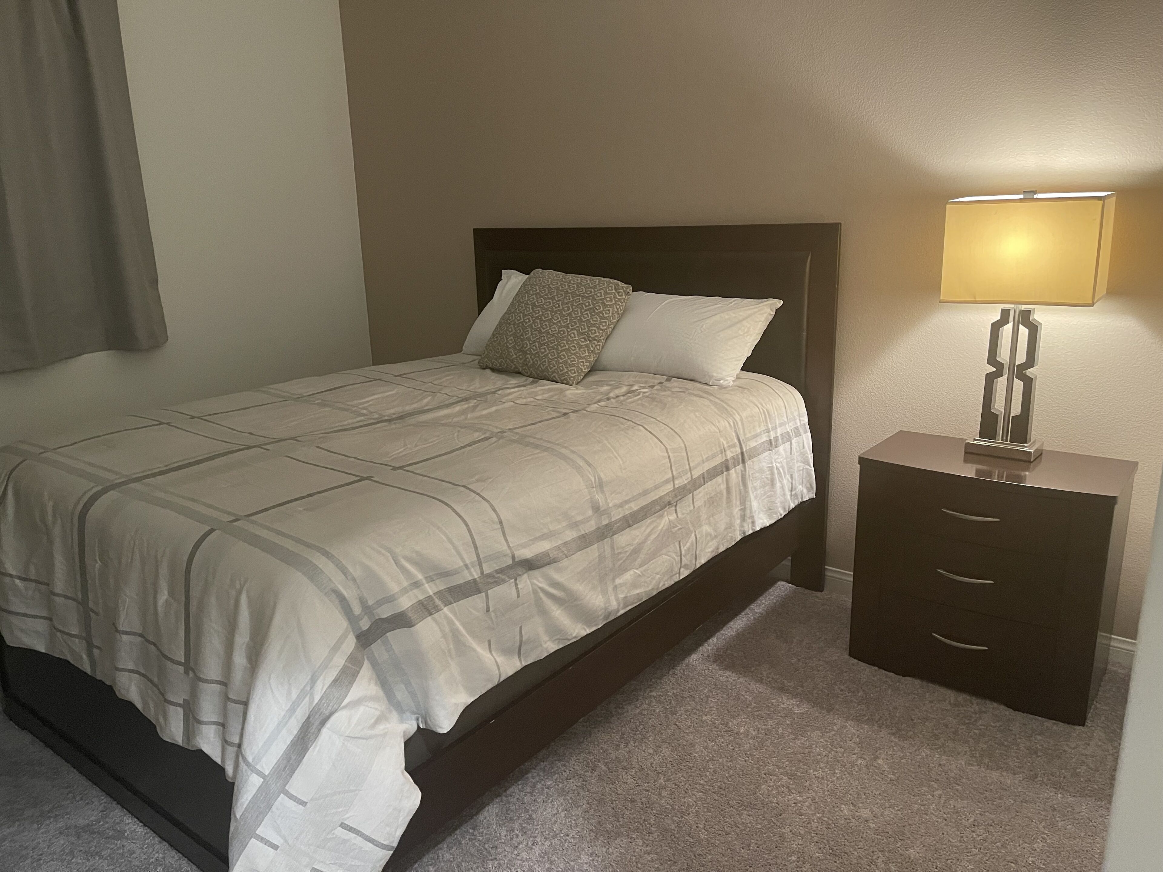 A neatly made bed with a patterned bedspread and pillows, a bedside table with a lamp, and a carpeted floor.