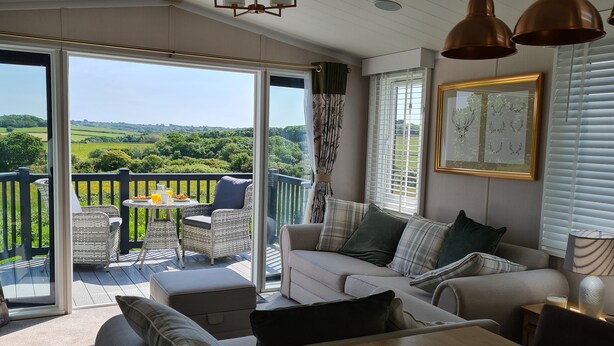 A cozy living room with a sofa, a coffee table, and a dining table. There is a balcony with wicker furniture and a view of the countryside.