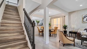 Una escalera con escalones de madera y barandillas negras, que conduce a un comedor con sillas y una mesa, y a una sala de estar con un sofá y una mesita de centro.
