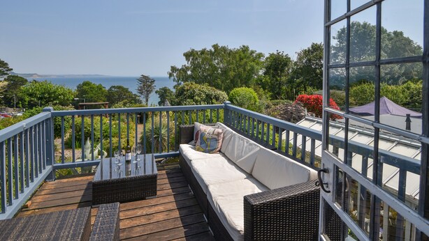 A balcony with a wicker sofa, a table with wine glasses, and a view of the sea. - Coram Cottage (Lyme Regis)