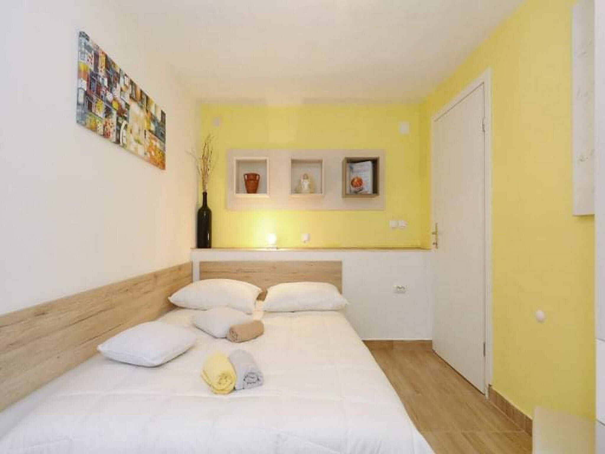 A bedroom with a bed, pillows, a bedside table, and a wall-mounted shelf with items.