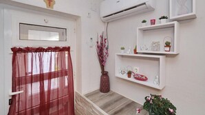 Un couloir avec un rideau rouge, une étagère blanche fixée au mur, ornée d’objets décoratifs, et un appareil de climatisation.