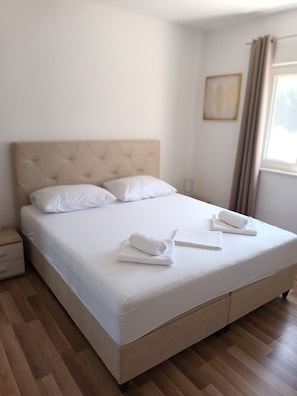 A neatly made bed with white linens and two rolled towels, a wooden nightstand, and a window with curtains. - Holiday Home Gabi - Three Bedroom Holiday Home with Terrace and Swimming pool (Šibenik)