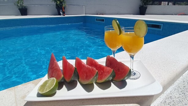 Une scène au bord de la piscine, avec un plateau blanc contenant des tranches de pastèque et un verre de jus d’orange agrémenté de rondelles de citron vert.