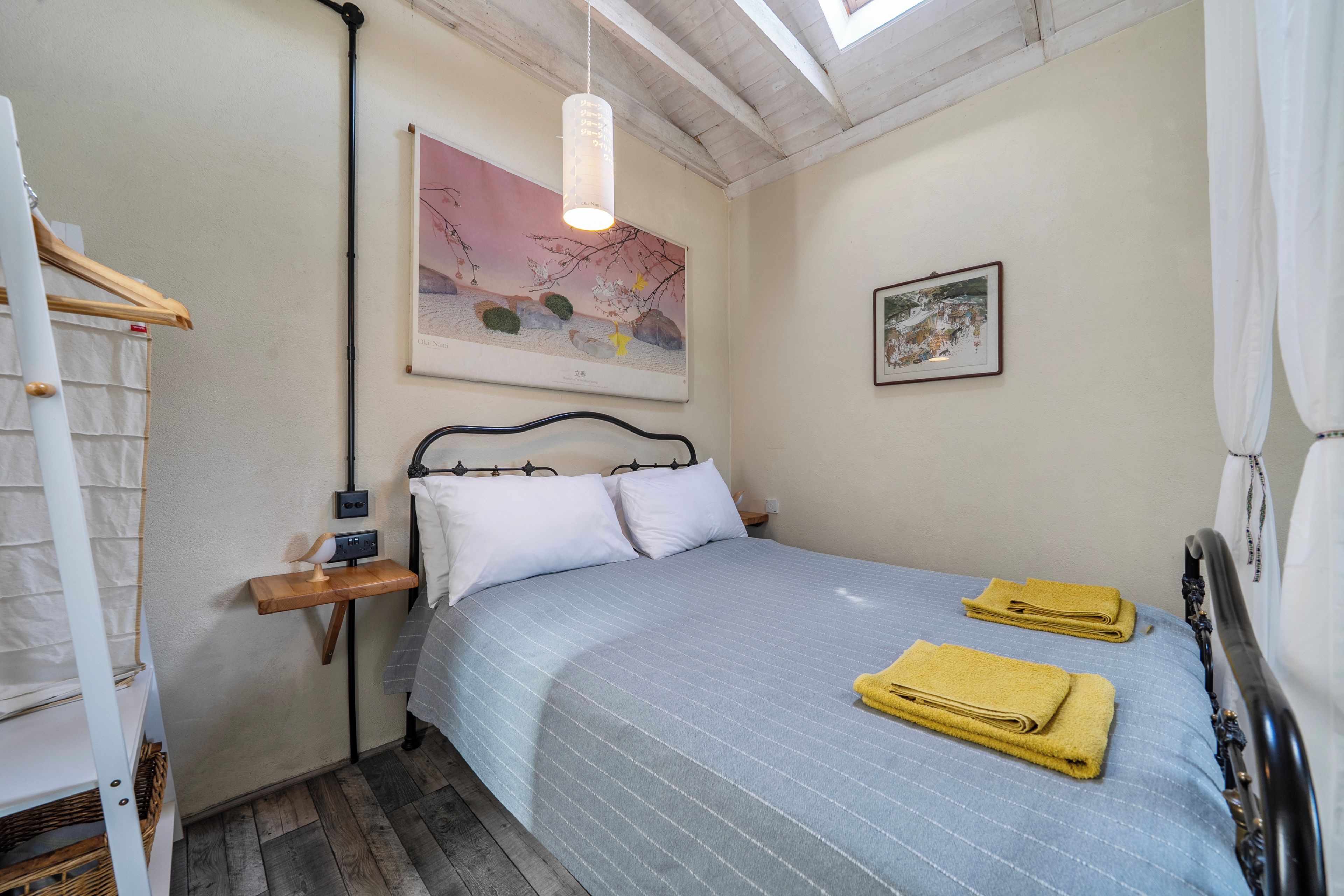 A bedroom with a bed, pillows, a bedside table, a hanging light fixture, a framed picture on the wall, and a window with curtains.
