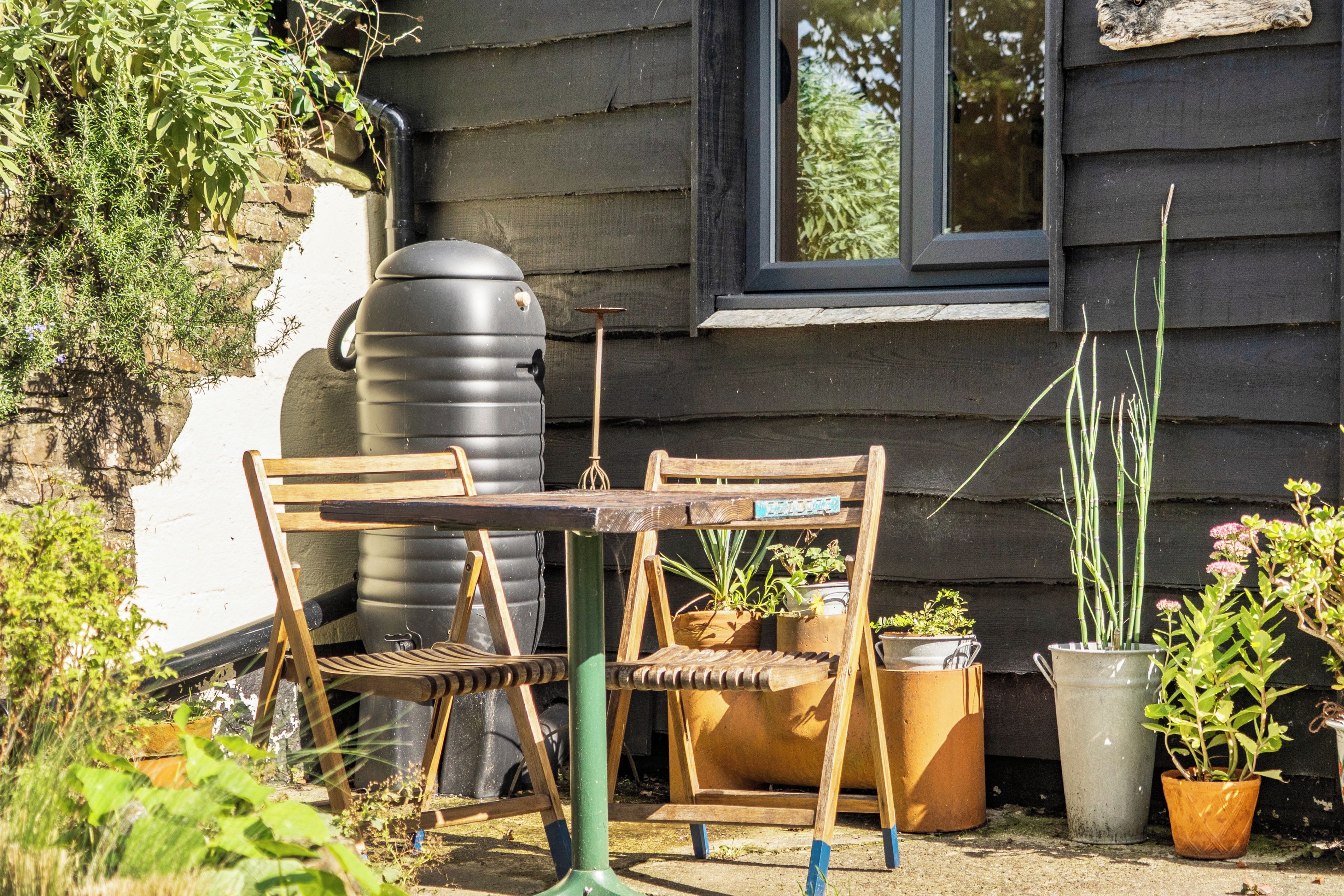 A rustic outdoor seating area with a wooden table and chairs, surrounded by potted plants and a large water tank.