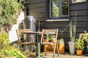 Un área de estar rústica al aire libre con una mesa y sillas de madera, rodeada de plantas en macetas y un depósito de agua grande.