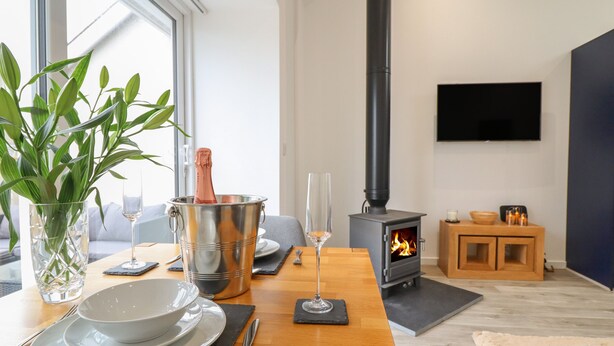 Ein modernes Wohnzimmer mit einem Holzesstisch, der für eine Mahlzeit gedeckt ist, einem Kamin, einem an der Wand befestigten Fernseher und einem großen Fenster, das natürliches Licht hereinlässt.