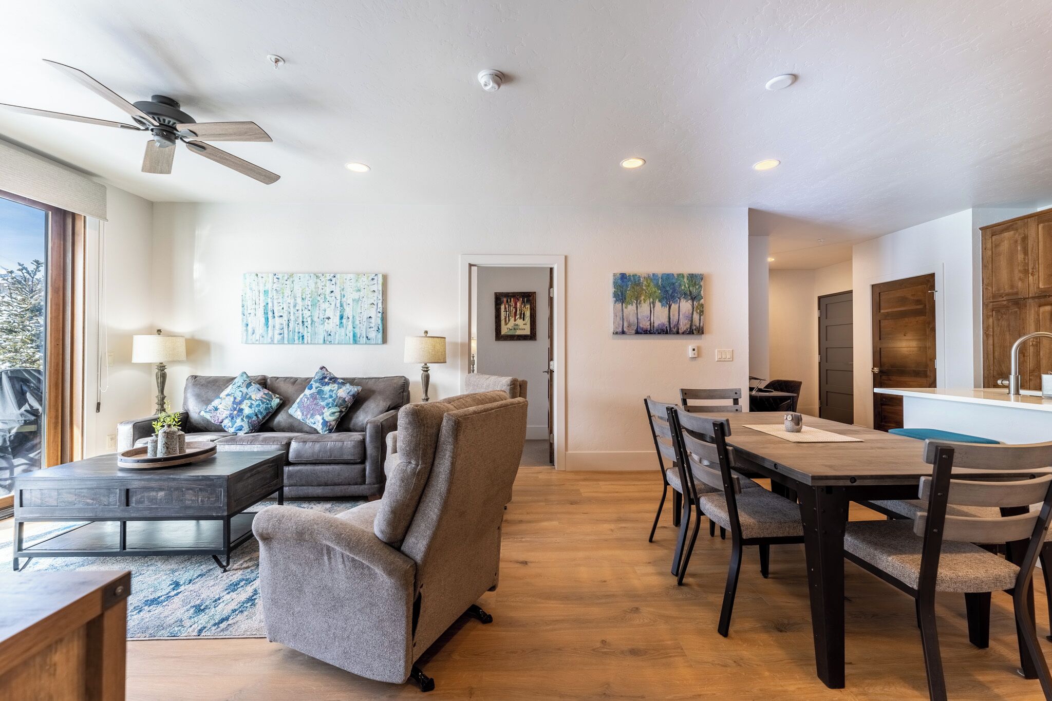 A modern living room with a sofa, coffee table, and dining table. There is a ceiling fan, recessed lighting, and artwork on the walls.