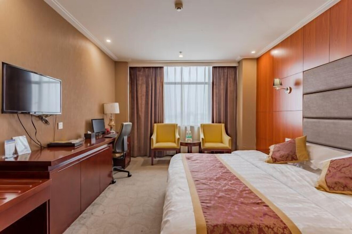 A hotel room with a large bed, a desk with a computer, two chairs, a TV, and a window with curtains.
