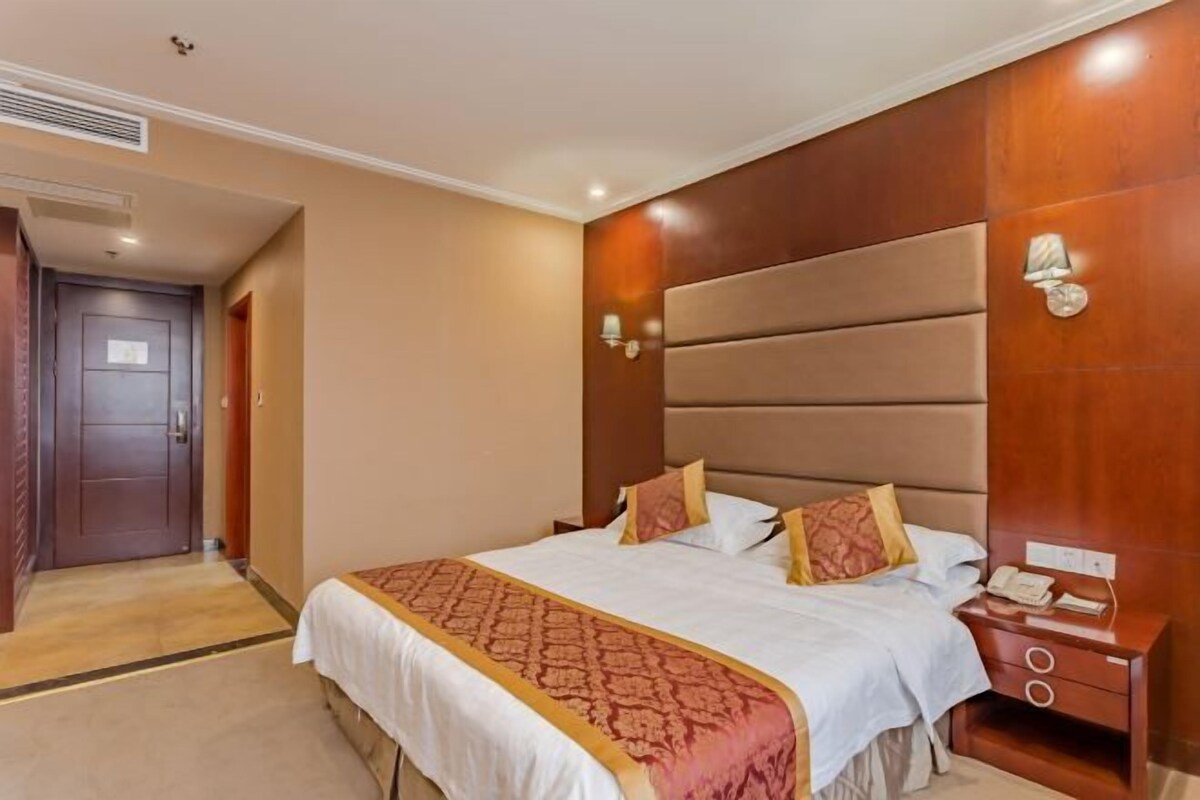 A hotel room with a large bed, a wooden headboard, a bedside table with a lamp, and a door in the background.