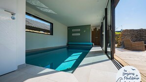 A modern indoor swimming pool area with a clear view of the outside, a wooden deck, and a stone wall. - La Longère de la Plage! Quiet, comfortable villa 300m from the beach! (Trégunc)