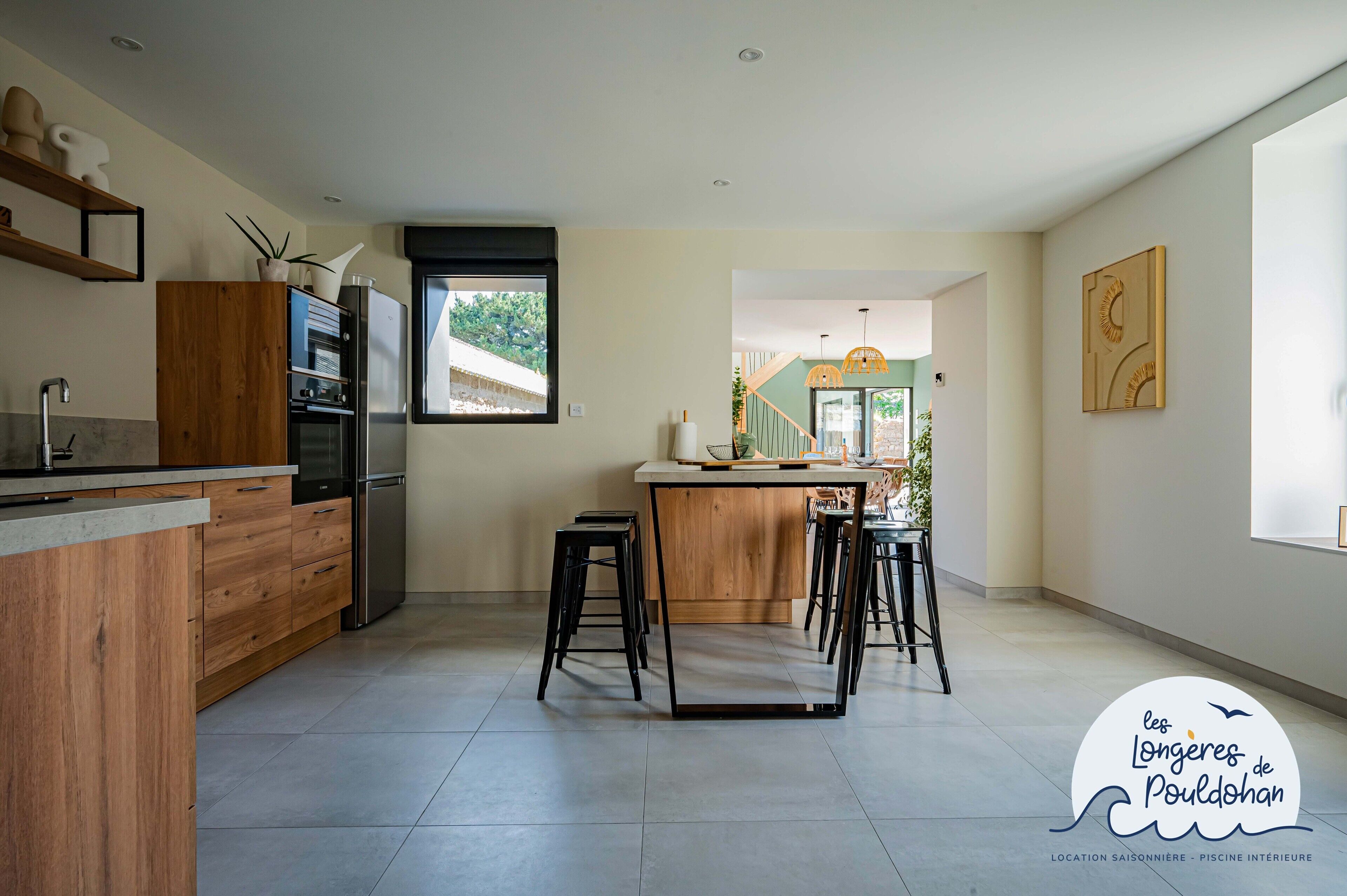 A modern kitchen with wooden cabinets, a central island with stools, and a view of the outdoors.