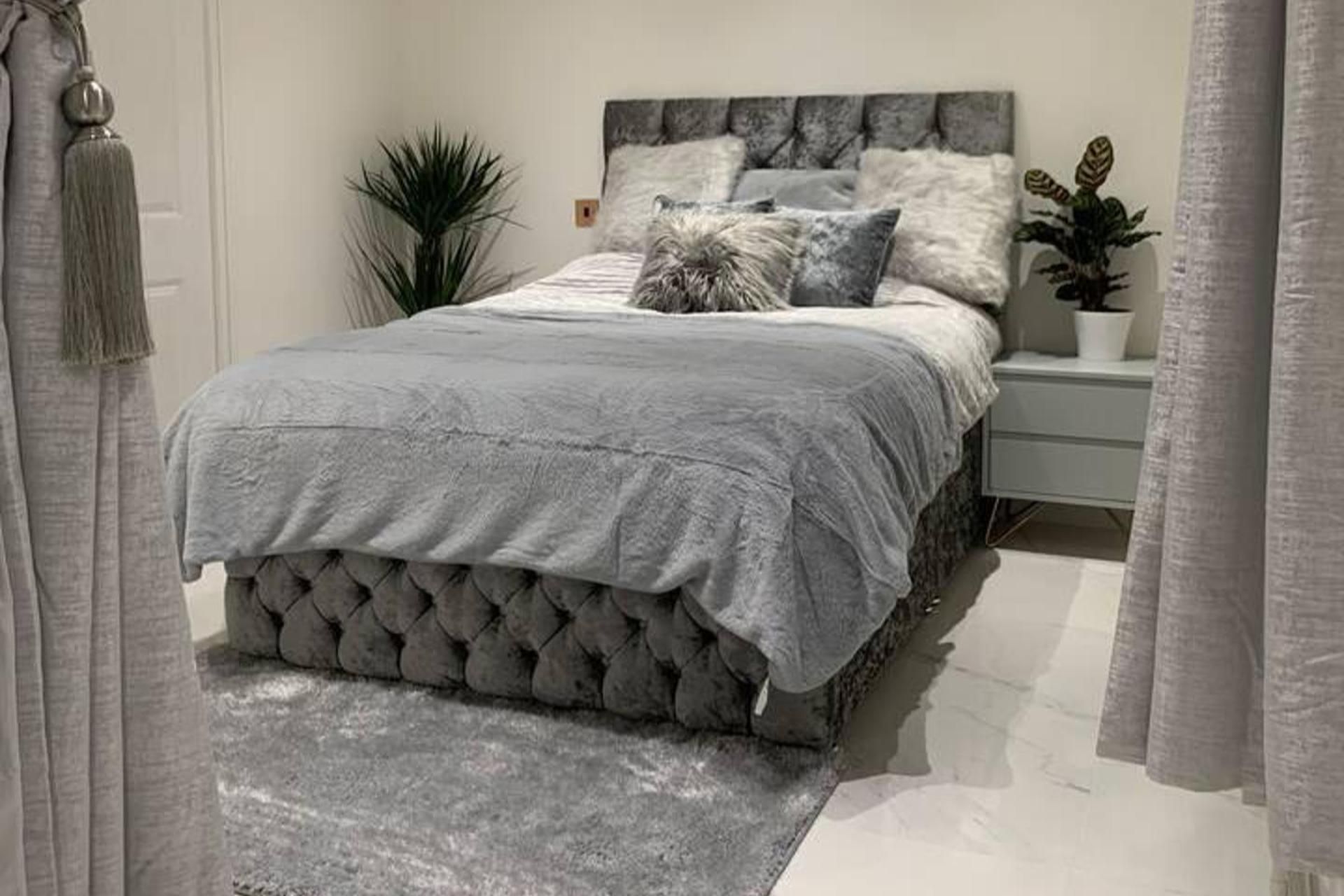 Une chambre à coucher avec une tête de lit capitonnée, une couvre-lit grise et une plante sur la table de chevet.