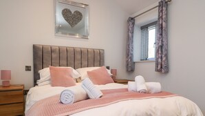 A neatly made bed with pink and white linens, a heart-shaped mirror on the wall, and a window with floral curtains. - Skirden View (Clitheroe)