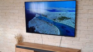 A modern living room with a flat-screen TV mounted on a textured wall, a wooden console table, and a patterned cushion on a white sofa. - Holiday Home Gabi (Bilice)