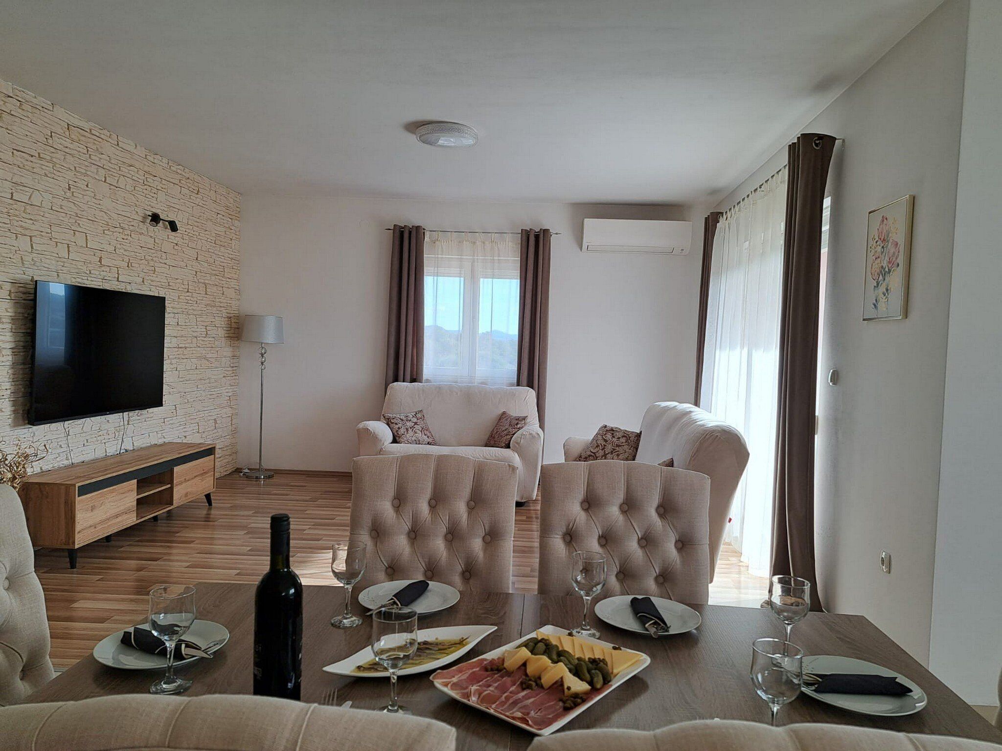 Uma sala de estar com televisão de tela plana, um rack de madeira, um conjunto de sofá, uma mesa de jantar com pratos e copos, e uma garrafa de vinho.