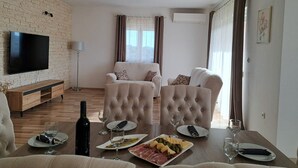A living room with a flat-screen TV, a wooden TV stand, a sofa set, a dining table with plates and glasses, and a wine bottle. - Holiday Home Gabi (Bilice)