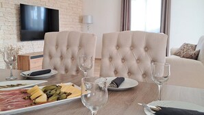 A dining area with a wooden table set for two, a flat-screen TV mounted on a brick wall, and a sofa in the background. - Holiday Home Gabi (Bilice)