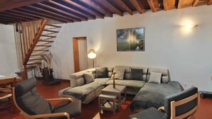 Ein gemütliches Wohnzimmer mit Holzboden, einem großen Ecksofa, zwei Holzstühlen, einem kleinen Tisch und einer Treppe, die zu einer oberen Ebene führt.