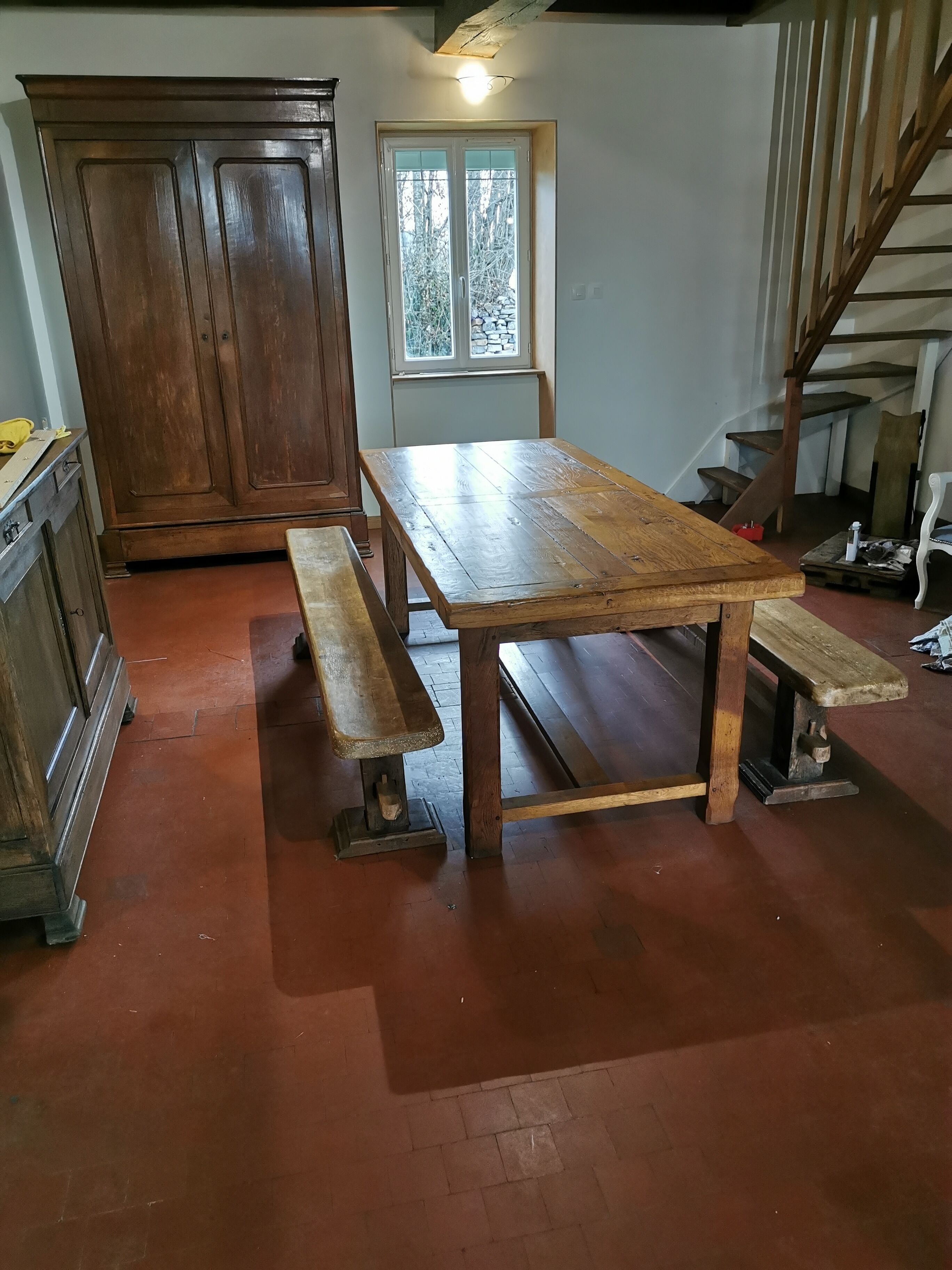 Een rustieke eetruimte met een houten tafel en banken, een grote kast en een trap.