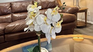 A living room with a brown leather sofa, a glass coffee table with white orchids, a lamp, and a blue abstract painting on the wall. - 5522 David & Jennie’s place 5 minutes to a community park (Houston)
