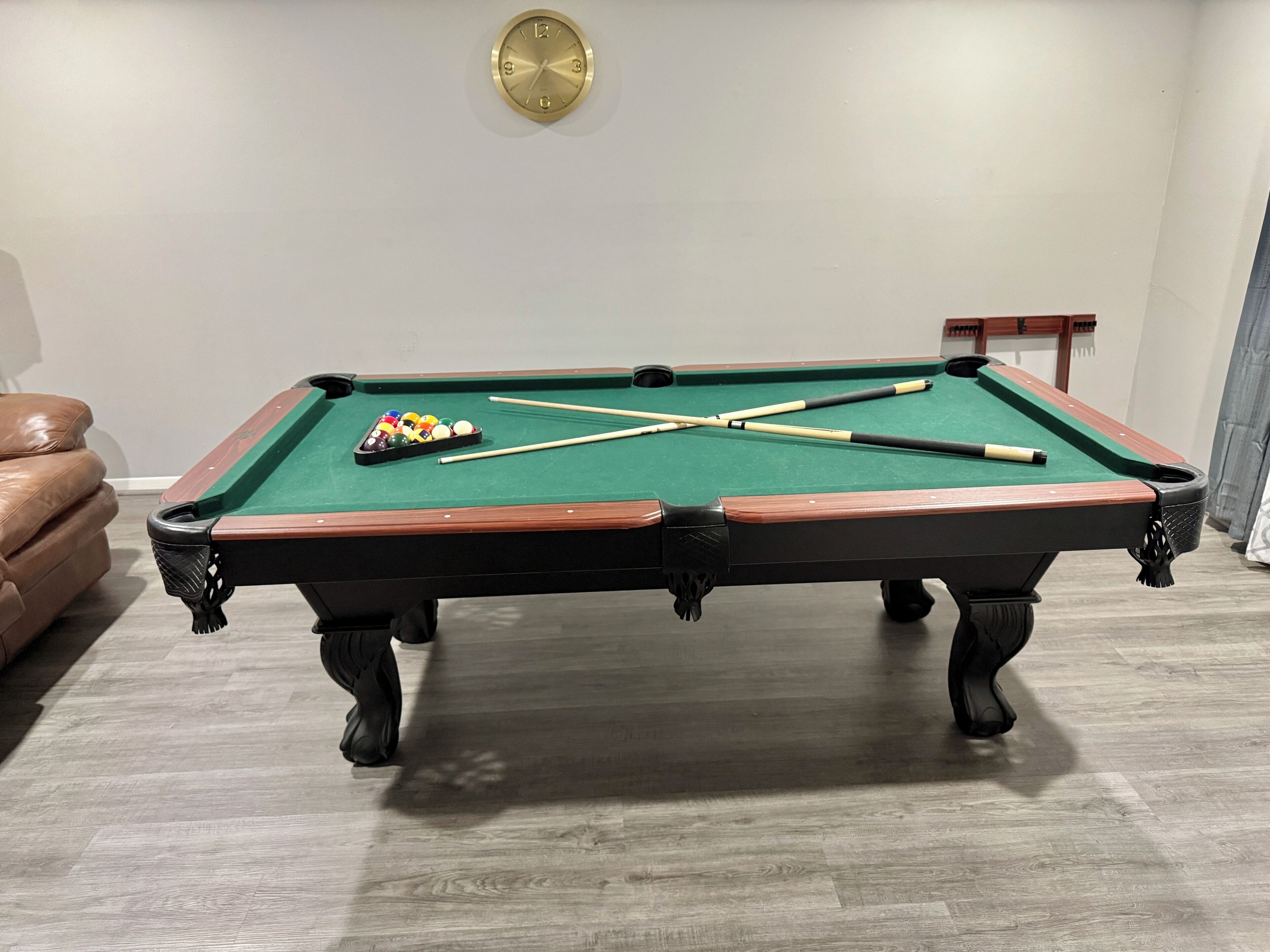 A pool table with balls and cues, a leather chair, and a clock on the wall.