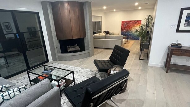 A modern living room with a grey sofa, a black ottoman, and a glass coffee table. There is a fireplace with a wooden mantel, a grey couch, and a colorful abstract painting on the wall. - Updated Modern House North Dallas! (Dallas)