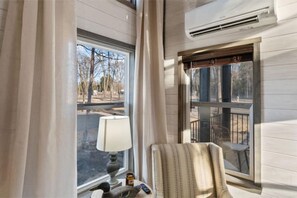 Relax in this inviting room with a comfortable chair and table, perfect for reading or unwinding, with a large window offering a view of a tree-lined street.