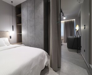 A modern hotel room with a large bed, a built-in wardrobe, a desk, and a seating area with a lamp.