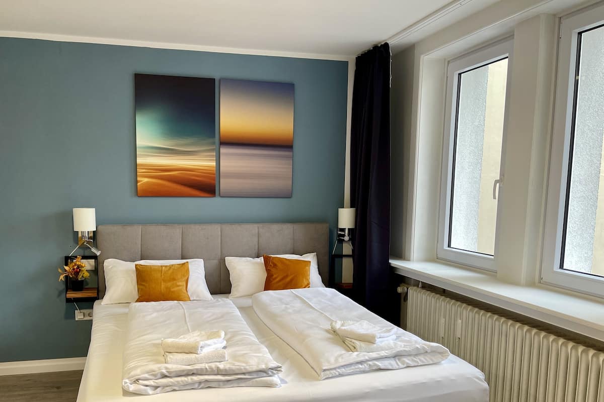 A hotel room with a large bed, two bedside tables, a nightstand, a window with curtains, and artwork on the wall.