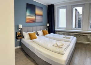 A double bed with white linens, a grey headboard, and two pillows. There is a bedside table with a lamp and a framed picture. The room has a large window with curtains, a radiator, and a view of buildings outside. - Aparthotel Neumünster City (Neumuenster)