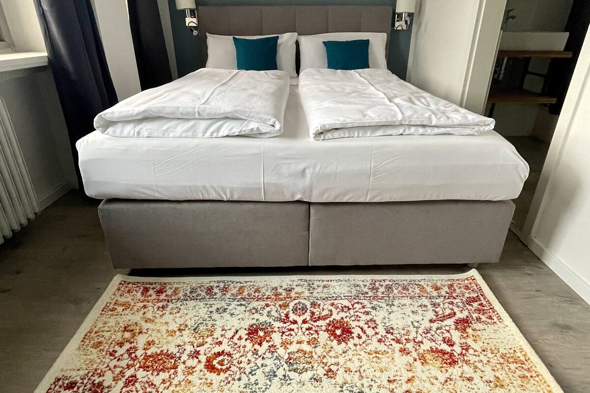 A double bed with white linens, a grey headboard, and a colorful rug on a wooden floor.
