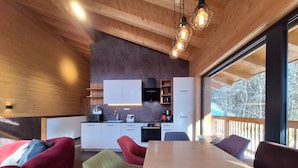 A modern kitchen with wooden cabinets, a dining area with a large table and colorful chairs, and a view of the outdoors through large windows. - Chalet Dinkel in Niedernsill 8-B (Niedernsill)