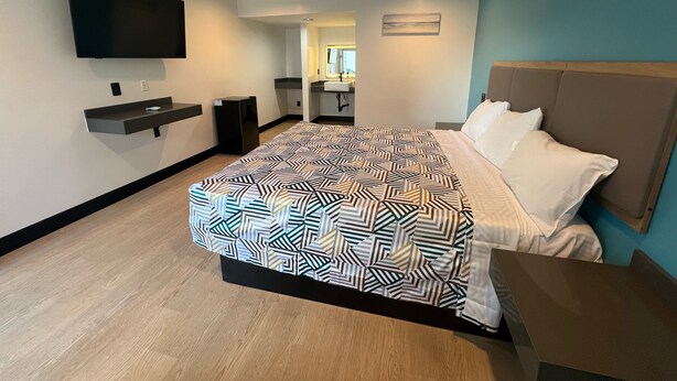 A modern hotel room with a large bed, a flat-screen TV mounted on the wall, a small desk with a chair, and a bathroom visible through an open door. - Arlo Motel (Inglewood)