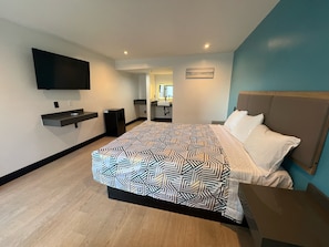 A modern hotel room with a large bed, a flat-screen TV mounted on the wall, a small desk with a chair, and a bathroom visible through an open door. - Arlo Motel (Inglewood)