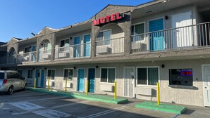 A motel with a sign reading "Motel" above the entrance. There are multiple doors with blue and white frames, and a parking area with marked spaces. - Arlo Motel (Inglewood)