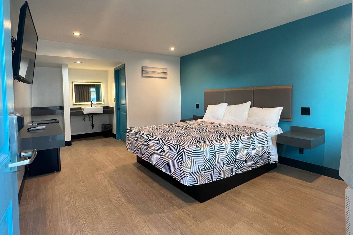Quarto de hotel moderno com uma cama grande, mesa de cabeceira, televisão, banheiro com pia e espelho, e piso de madeira.