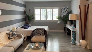 Un espace de vie compact comprenant un canapé, deux fauteuils, une petite table avec une bougie, un lit, une fenêtre avec des stores et une lampe décorative.