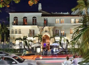 Classic two-story building with an arched entrance and upper balcony, set amidst a fenced area with palm trees.