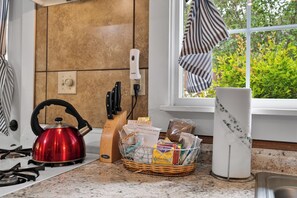 A kitchen with a red kettle, a knife block, a basket of groceries, and a window with a view of greenery.