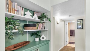 Un couloir avec un meuble vert, des étagères blanches ornées de livres et de plantes, et une porte donnant sur une salle de bain.