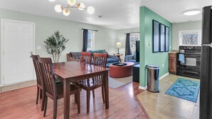 Une cuisine moderne avec un réfrigérateur noir, une cuisinière et un évier. On y trouve un coin repas avec une table et des chaises en bois. Les murs sont peints en vert clair, et il y a une fenêtre avec des rideaux.