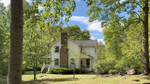 A two-story house with a stone chimney, surrounded by trees and a grassy yard. - Hightop Hideaway, a cozy secluded mountain retreat. Pet-friendly, Shenandoah NP. (Stanardsville)