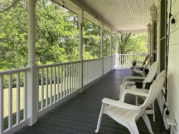 A covered porch with white railings and wooden flooring, featuring two white plastic chairs and a table.