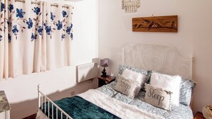 Un dormitorio con cama, mesita de noche, lámpara, radiador, ventana con cortinas y una obra de arte en la pared.